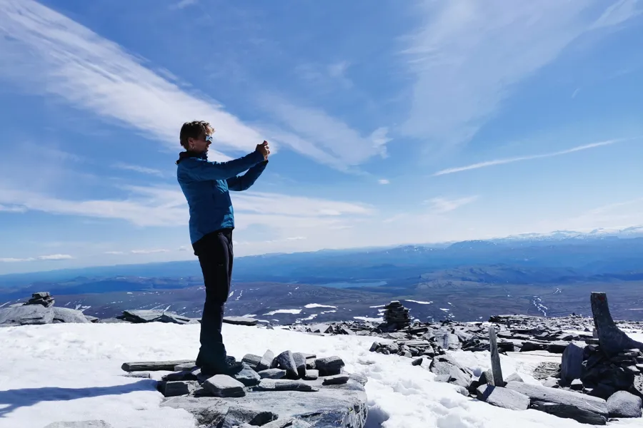 Dame står på en fjelltopp i sol en i en stor fjellheim og fotograferer landskapet