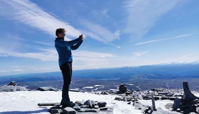Dame står på en fjelltopp i sol en i en stor fjellheim og fotograferer landskapet