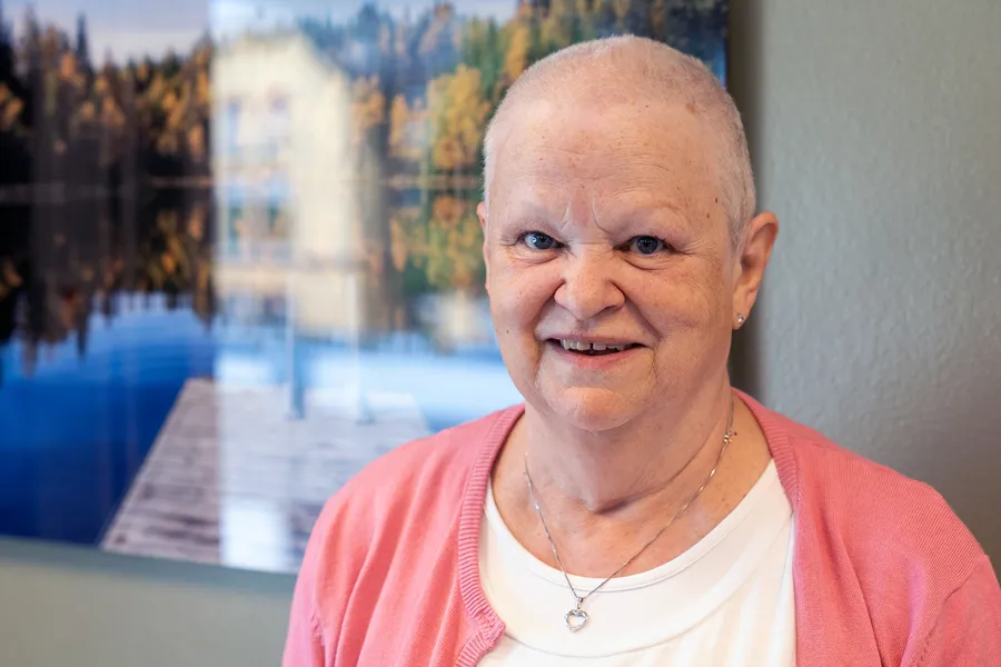 En blid kvinnelig kreftpasient med rosa genser og nesten ikke hår står foran fotokunst på et venterom på et kreftsenter