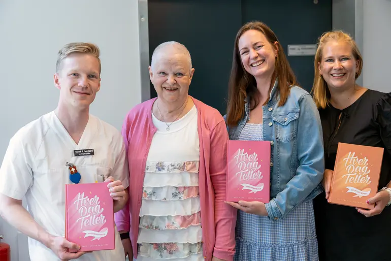 Fire smilende fjes. En mannlig fagsykepleier i hvitt, en eldre dame i rosa med kortklipt hår (kreftpasient) og to blide damer