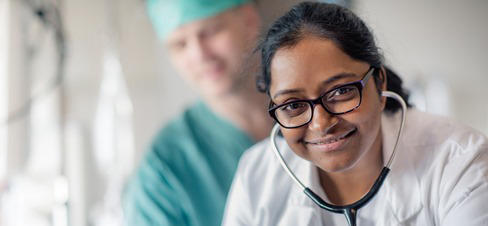En lege med stetoskop smiler, i bakgrunnen ser man en kirurg.