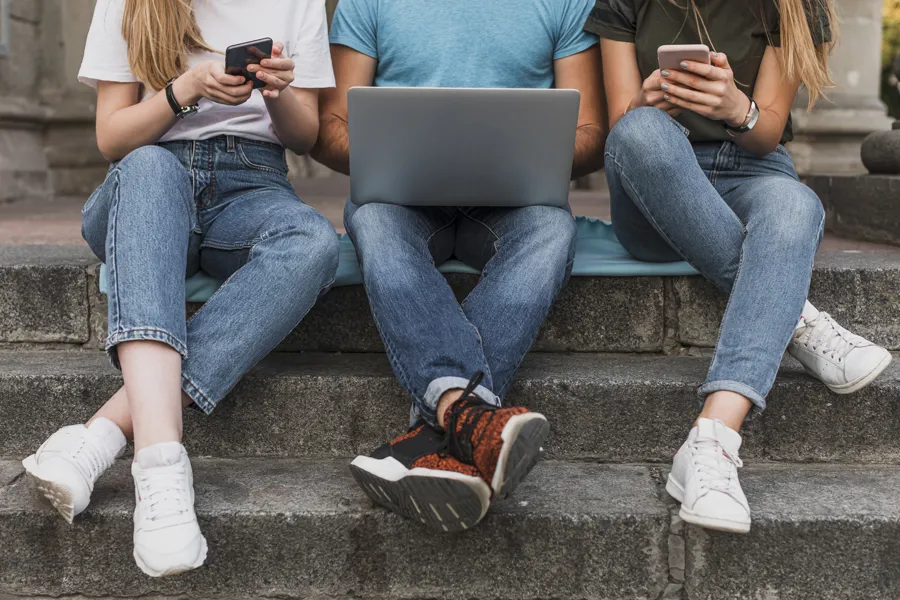 Tre ungdommer sitter ved siden av hverandre i en trapp og ser på hver sin mobil- og PC-skjerm