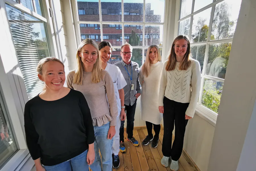 Seks personer står samlet foran et vindu